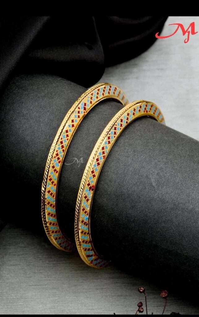 A close-up image of two intricately designed gold bangles featuring colorful patterns of red and blue gemstones, resting on a dark surface.